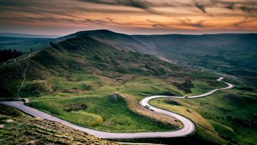 The winding road at Mam Tor. Mam Tor is green and grassy and the sky is red as if the sun is just starting to set. 