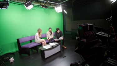 Three students in front of a green screen ready to record a news programme. 