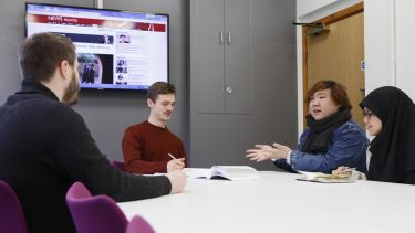A group of politics students have a discussion around a table. BBC News is on in the background. 
