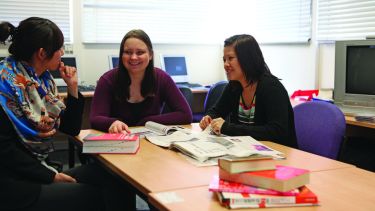 Three female postgraduate students sat at a table looking at a newspaper