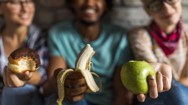 Close up of happy friends holding banana, apple and a donut.