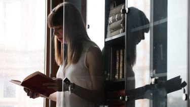 A history student standing next to a bookcase reading a book. 