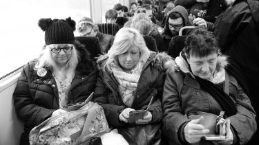 Black and white image of three women sat on a crowded train, all checking their phones