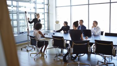 A group of people sitting around a boardroom table in a business meeting. One woman is presenting to the group.