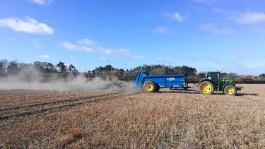 A tractor pulls a trailer across a field of dry grass spreading rock dust in a long cloud