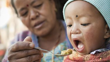 Outdoor image at day time of Asian woman feeding food to her grandson. The little child is opening his mouth wide to eat his lunch fed by his grandmother. Two people, Head and shoulder, horizontal composition and selective focus.