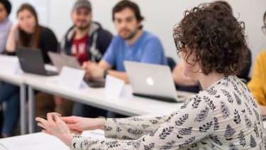 Felicity Matthews is in the foreground of the image, speaking to a group of students sat at desks with laptops.