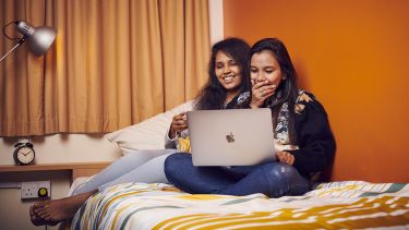 Photo of two students sat on a bed laughing at a laptop screen