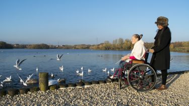 A wheelchair user and her carer visit a park, they are next to a pond with birds.