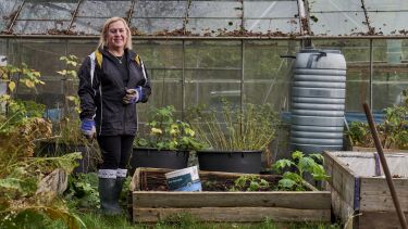 A woman is stood in an allotment next to a raised bed. There is a greenhouse in the background and several tubs of plants growing