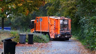 An orange garbage truck driving down a road where the black bins have been put out.