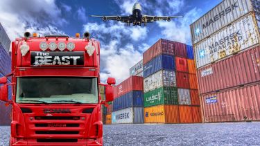 A red lorry named 'The Beast' in the foreground. Behind it are stacks of shipping containers. In the sky is an aeroplane.