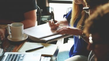 People sat round a table working on a project. On the table there is a mug, notebooks, pens and a laptop.