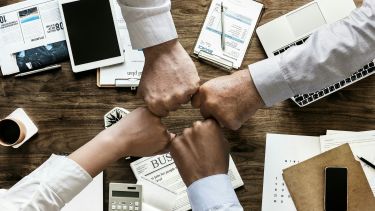 Four fists put in the centre of a circle on top of a desk covered in notebooks, documents, stationery and handheld electrical devices