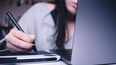 Woman sat at laptop drawing on tablet with stylus