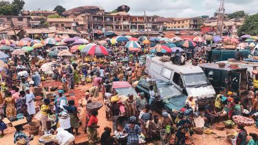 A photograph of a busy area in an African city. There are dozens of market stalls under umbrellas. There are several vehicles parked around the edge.