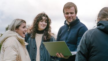 A man holding a laptop outside, showing the screen to a group of students