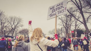 Picture of a protest with a woman holding a sign reading "we are better than this"
