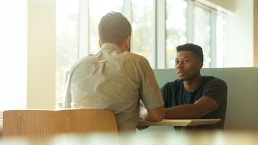 Two people having a seated conversation in front of a window. 