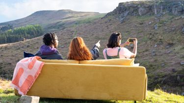Three prospective students sit on a yellow sofa on a hill overlooking the picturesque Peak District