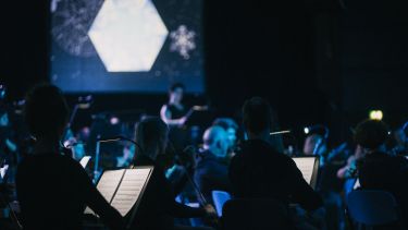 An orchestra in pale blue light perform in the Octagon, imagery of snow is projected onto a large screen in the background