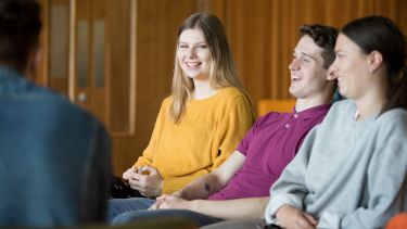 Three students sit against a wooden backdrop, laughing.