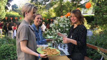 Three students at a party in a garden. Two carrying plates of food and presenting to the third.
