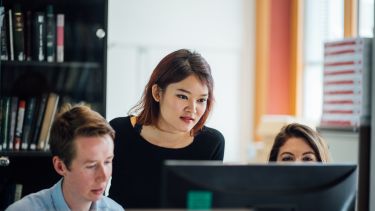 Image of three students sitting together at a desk and looking at a computer.