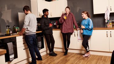 Photo of diverse group of students stood in a kitchen talking and drinking 