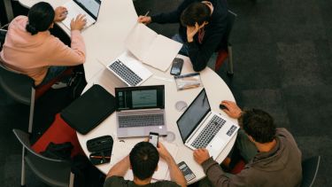 A group of students working in the Diamond with laptops, huddles around a circular table.