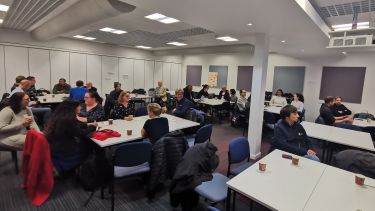 Insigneo General Assembly event view of people sitting talking at tables in a university workroom