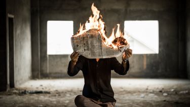 A person sat on a chair holding a flaming newspaper.