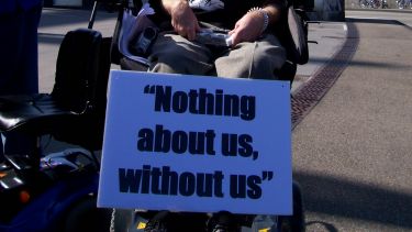An image of a disabled activist holding a sign reading 'Nothing about us without us' by Elisney is licensed under CC BY-NC-ND 2.0.