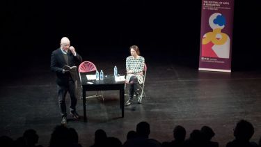 A book reading taking place, with one individual sat on a stage and another stood, holding an open book