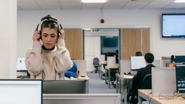 A student stands in a computer room and is putting a pair of headphones on 