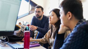 Students sit in front of a computer, being guided by a tutor