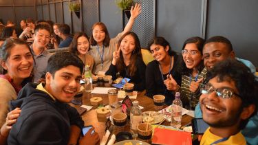A group of smiling students gathered around a table full of drinks in a cafe