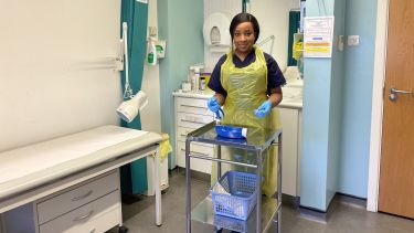 Racquel, a general practice nurse wears an apron and gloves in front of a medical trolley