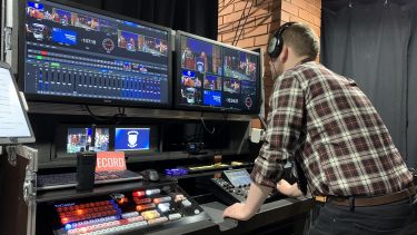A person with their back to the camera, looking at a screen with several different camera angles of video on it, and a mixing desk in front of them