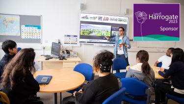 A picture of a teacher stood in front of 6 students sat at desks in a classroom demonstrating an English language teaching concept with the aid of a smart board behind him
