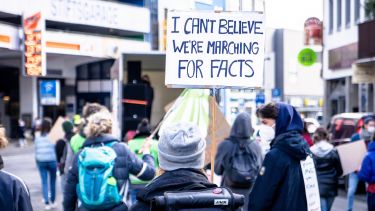 A protest on a street with a woman in the foreground carrying a sign that says, 'I can't believe we're marching for facts'