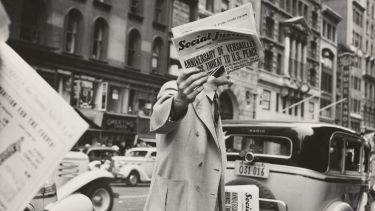 A man dressed in early twentieth century clothing, stood on a street surrounded by old motor vehicles, holds a newspaper up