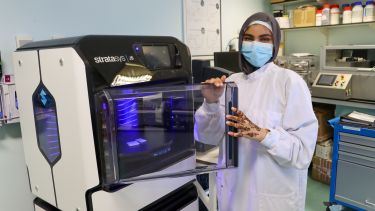 Aliza wearing a lab coat and face mask using a high tech 3D printer