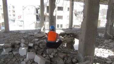 An academic kneeling down to inspect rubble inside a damaged building in Syria