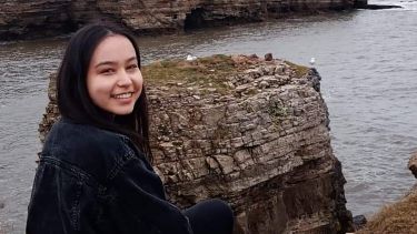 Picture of Yoana sitting outside on a grey and cloudy day, on the edge of a coastline with a view of birds nesting on isolated rocks and cliff faces. 