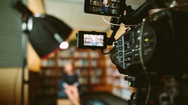 a close-up of a video camera pointed at a woman. The woman is sitting in a chair in front of a large bookshelf and appears to be giving an interview.