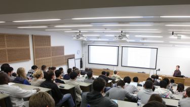 Backs of heads of students in a tiered lecture theatre with two screens at front and presenter stood at lectern box