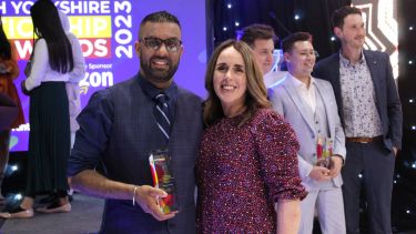 A man in a suit holds up his award to the camera, with his arm round a woman wearing a purple dress.