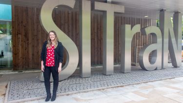 A woman with long blonde hair wears a black cardigan and red top. She stands in front of a metal set of letters spelling 'SITraN'