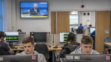 Two boys in grey hoodies, looking down at computer screens. They are in a large room surrounded by other people sat at computers, as well as a tv screen showing a sky news broadcast in the background.